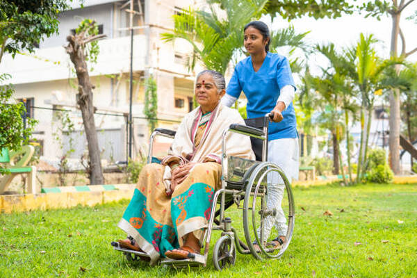 wide shot of Nurse taking senior woman on walk while on wheelchair at hospital garden - concept of caretaker, disability and healthcare.